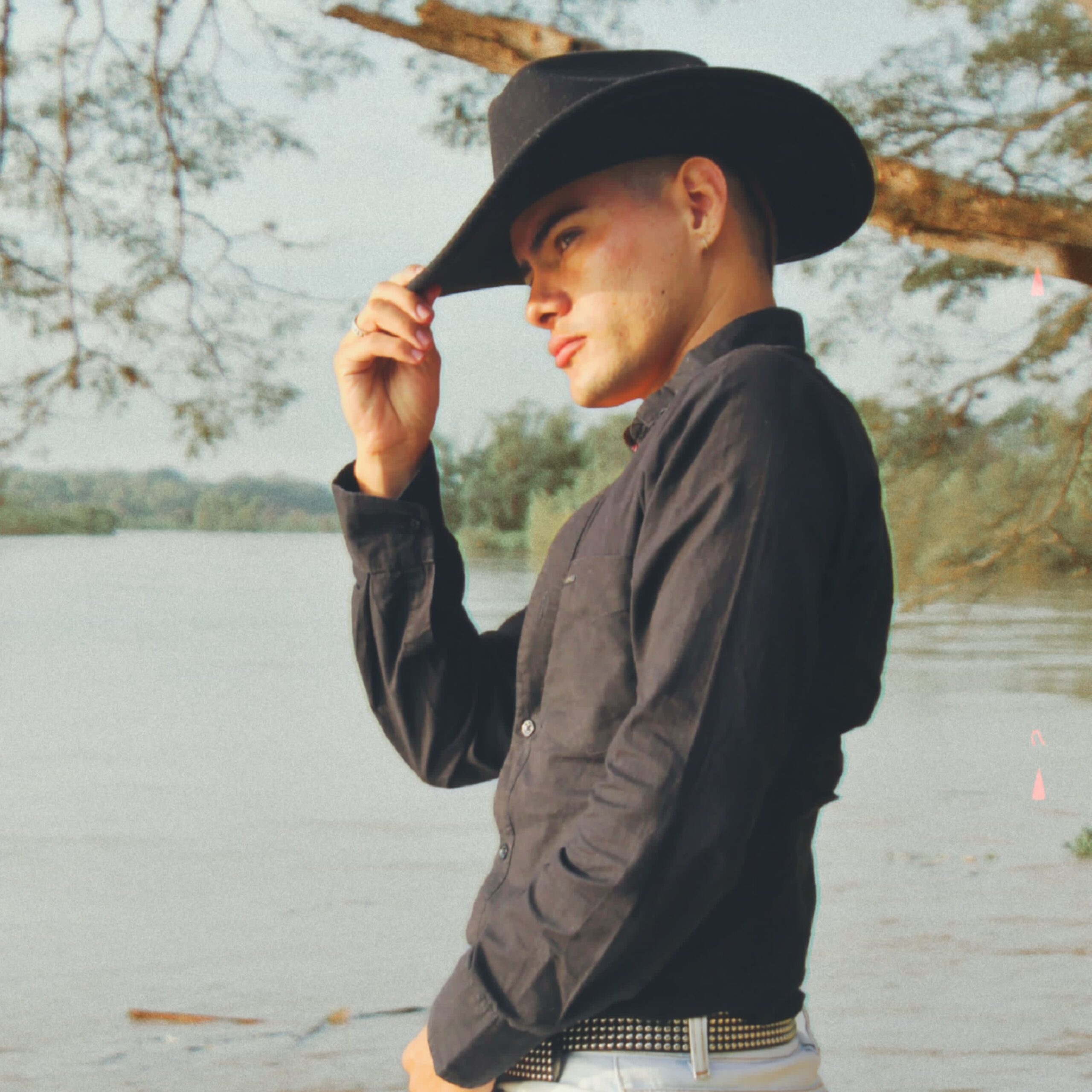 Liam Kai is facing left while pulling a black cowboy hat over down to shade his eyes. He is wearing a black dress shirt, white jeans and is standing in front of a wide river
